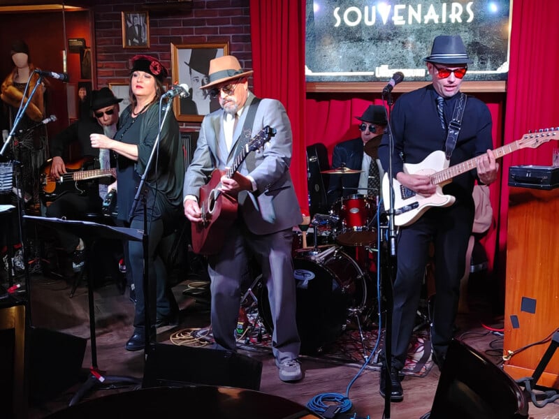 A band performs in a dimly lit venue. Four members are on stage: a woman singing, two men playing guitars, and a drummer in the back. They wear dark hats and sunglasses. A neon sign reading "SOUVENIRS" glows behind them.