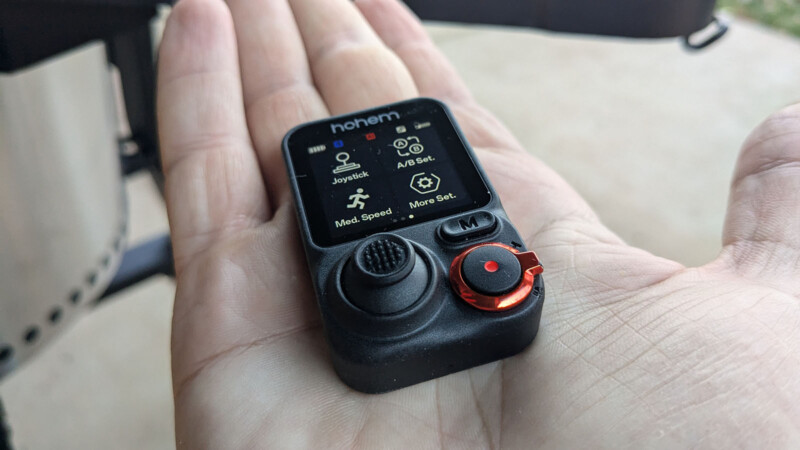 A close-up of a hand holding a small black remote control device. The device has a display screen, labeled "hohem," showing icons and text for joystick, balance, speed, and more settings. It features a joystick and red dial.