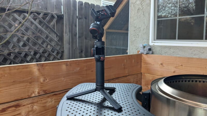 A camera stabilizer positioned on a table outdoors. The stabilizer is black with red accents and has a tripod base. It is set against a wooden fence and a building with a window. A small, gray plush toy is visible near the window.