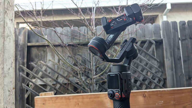 A handheld gimbal stabilizer with a camera mount is positioned outdoors against a wooden fence and leafless trees. The gimbal appears to be powered on with its screen visible. The scene is set in a backyard or garden area.