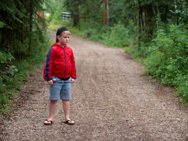 A young boy stands on a gravel path in a forest. He wears a red jacket, blue shorts, flip-flops, and a black cap. The path is surrounded by lush greenery and trees. The boy looks to the side with a thoughtful expression.