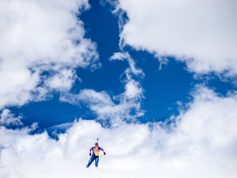 A skier dressed in vibrant gear speeds downhill on a snowy incline beneath a brilliant blue sky dotted with fluffy white clouds.