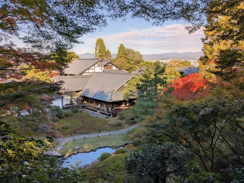 A serene Japanese garden with a traditional building surrounded by vibrant autumn foliage. The scene includes red, orange, and green trees, a small pond, and rolling hills under a partly cloudy sky.