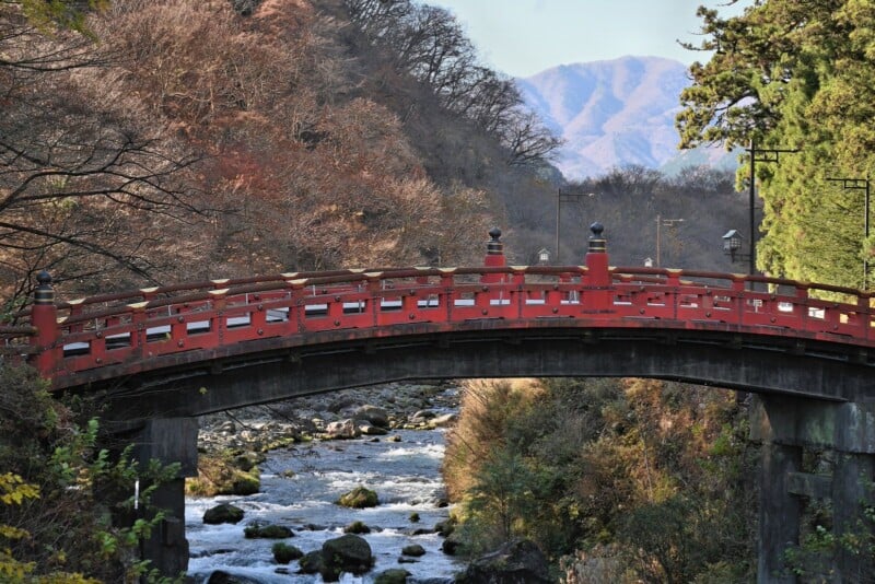 A traditional red wooden bridge arches over a flowing river, surrounded by trees with autumn foliage. In the background, hazy mountains rise under a clear blue sky.