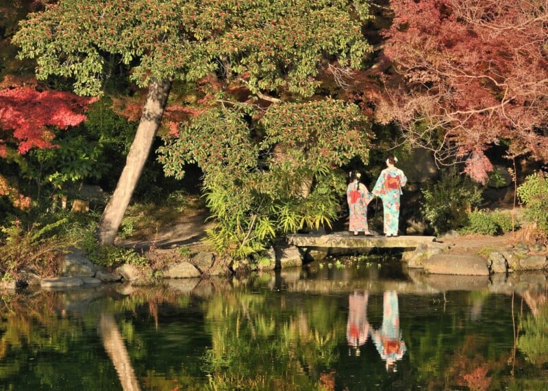 Two people in colorful traditional clothing stand beside a serene pond. They are surrounded by vibrant autumn foliage, with red and green trees reflected in the water. A small stone bridge is visible under the tree canopy.