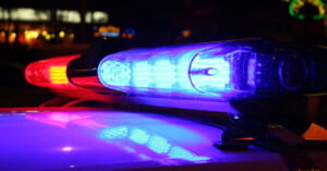 Close-up of a police car light bar at night, with blue and red lights illuminated, reflecting on the vehicle's surface. Blurred lights are visible in the background.