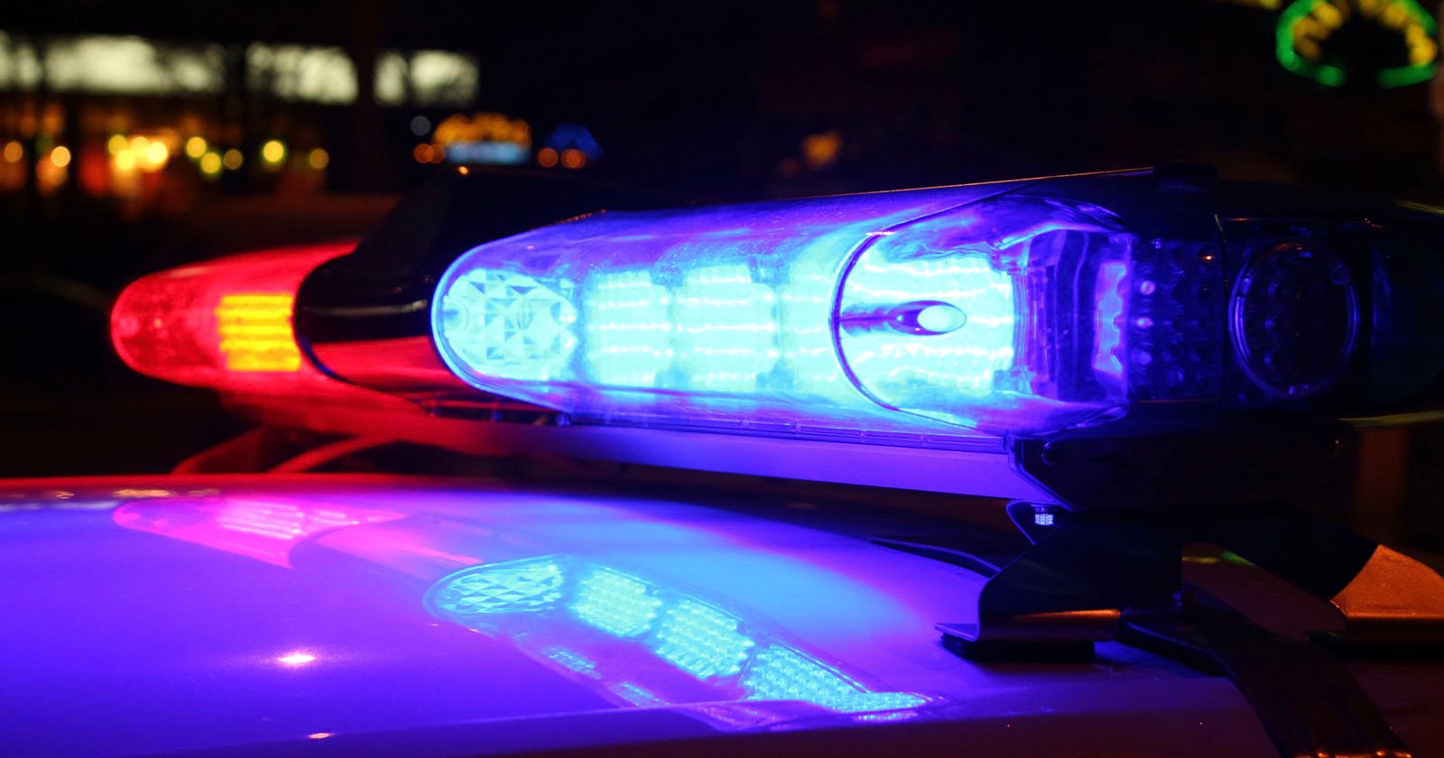 Close-up of a police car light bar at night, with blue and red lights illuminated, reflecting on the vehicle's surface. Blurred lights are visible in the background.