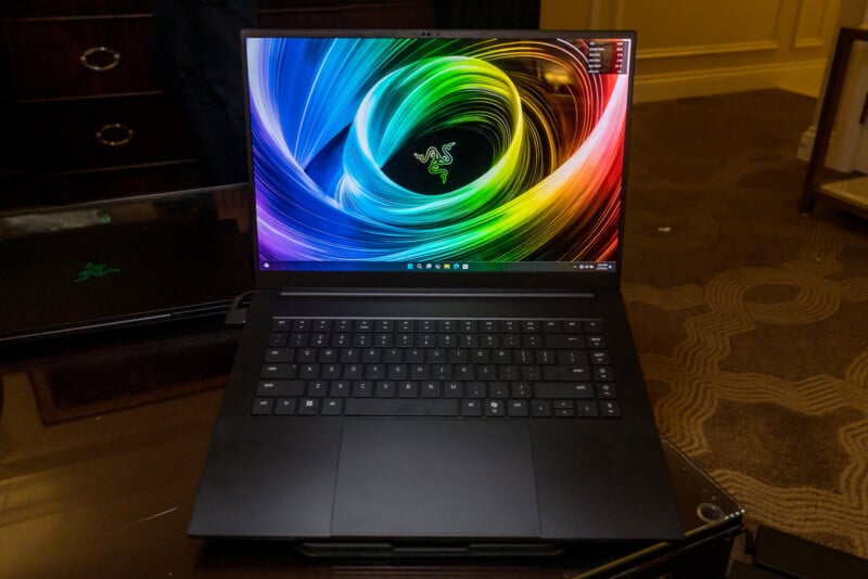 A sleek black laptop on a glass table with a colorful spiral design on the screen. The background features a carpeted floor and a dresser.