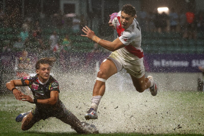 A rugby athlete clad in a white and red outfit leaps to evade a tackle on a waterlogged field, sending droplets scattering. Another player in a dark outfit reaches out in an attempt to tackle him. Spectators and stadium lights can be seen in the background.