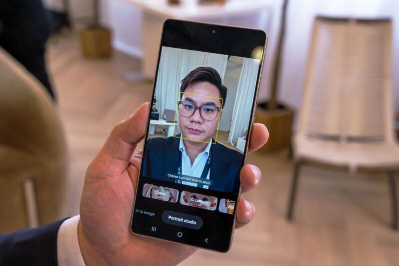 A person holding a smartphone displaying a facial recognition interface. The screen shows a person wearing glasses within a yellow square. Background features blurred indoor elements like chairs and flooring.