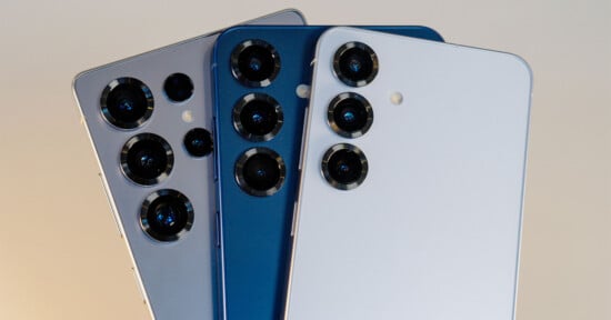 Three smartphones are displayed, each with three rear cameras and different colors: silver, dark blue, and white. The phones are angled to highlight their camera designs against a neutral background.