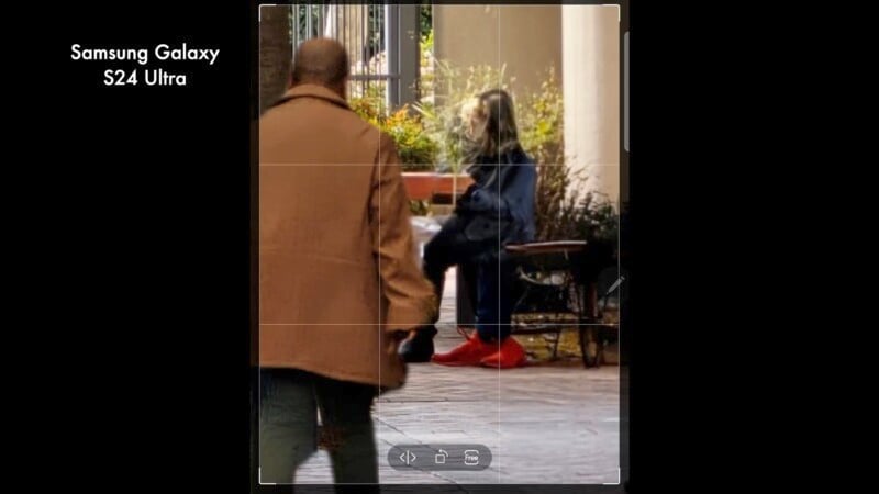 A person in a brown coat walks toward another person sitting on a bench wearing red shoes and dark clothing. It's a street scene with plants in the background.