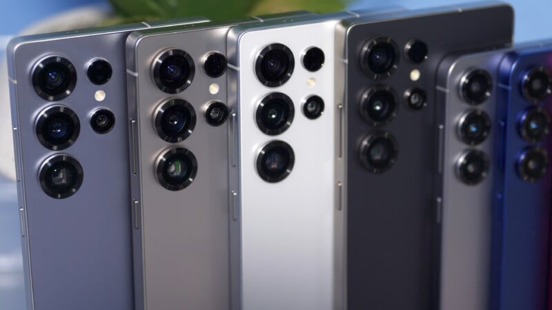 Several smartphones with triple-camera setups are displayed in a row, featuring different colors such as silver, gray, and black. The focus is on the back of the phones, showcasing the camera designs.