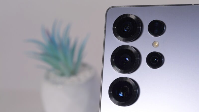 Close-up of a smartphone's rear camera module featuring multiple lenses and a flash. A blurred plant in a white pot is visible in the background.