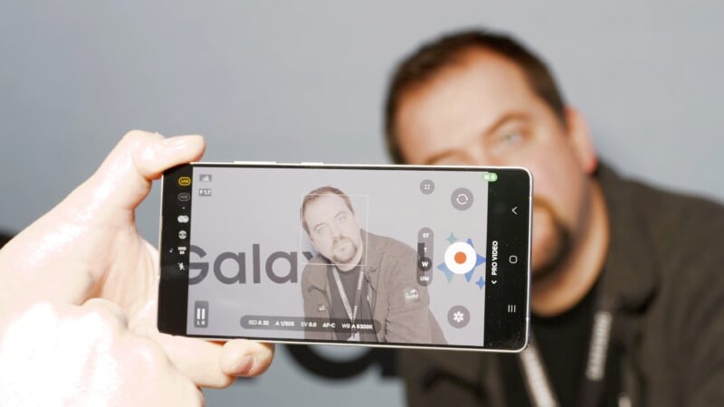 A person is holding a smartphone, capturing a photo of a man in the background. The phone screen displays the camera interface, showing a black-and-white image of the man, who is wearing a lanyard and a beard. The word "Galaxy" is partially visible.