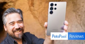 A person smiling holds up a silver Samsung smartphone with multiple cameras. The background is a blurred natural setting. In the foreground, there is text reading "PetaPixel Reviews.