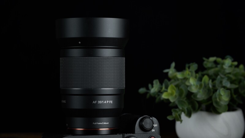 A black camera lens labeled "AF 35/1.4 FE" mounted on a camera against a dark background. A small potted plant with green leaves is blurred in the background, adding a touch of color.