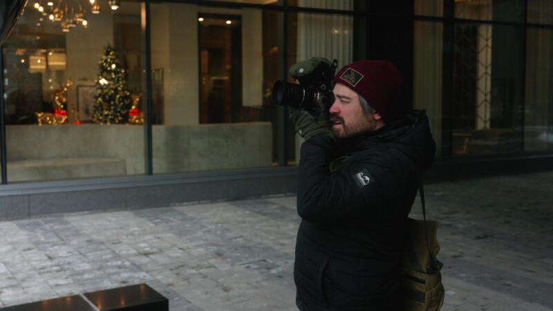 A person in a winter jacket and red beanie takes photos with a camera on a city street. In the background, a window display features a decorated Christmas tree and festive lights.