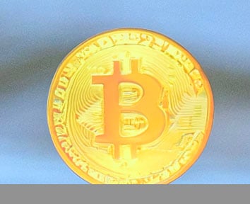 A close-up of a shiny gold coin with the Bitcoin symbol embossed on it, set against a blue background. The coin features intricate designs and text around the edges.