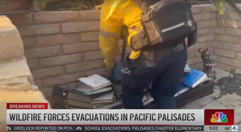 A person in a yellow jacket and backpack is stacking binders and books outside next to a brick wall. The news headline reads, "Wildfire forces evacuations in Pacific Palisades.