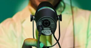 Close-up of a person holding a Shure MV7 microphone, showing the input connections at the back. The person is wearing a light-colored shirt, and the background is a soft green.