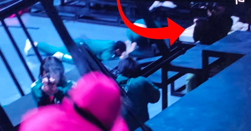 Several people in green uniforms are crawling on the floor under a structure, while a person in a pink outfit with a mask observes them. A large red arrow is pointing towards one of the individuals in green. The scene is dimly lit.