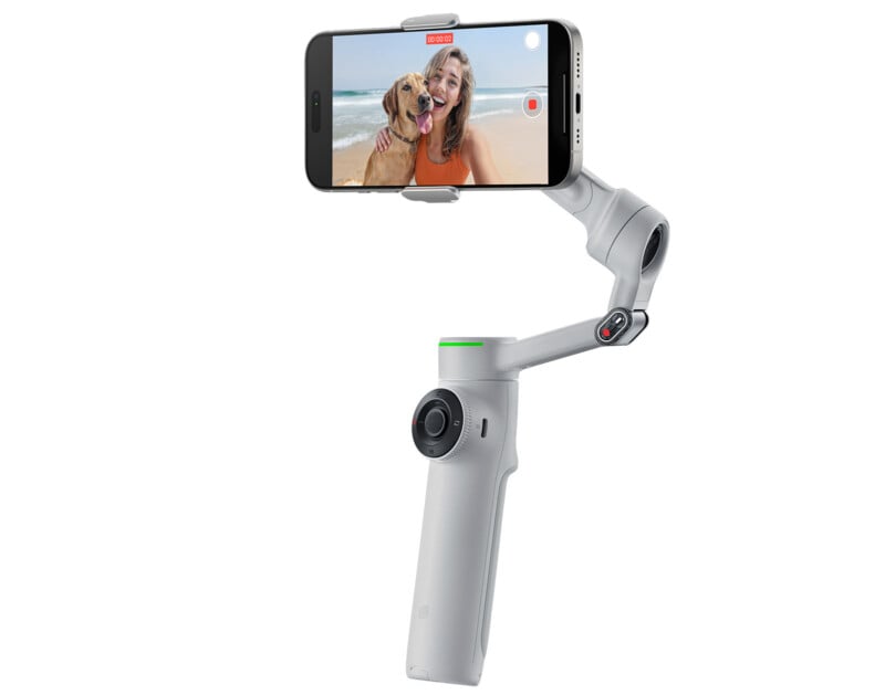 A smartphone mounted on a white gimbal records a video. The phone screen shows a woman at the beach hugging a dog, both smiling. The gimbal has control buttons and an ergonomic grip.