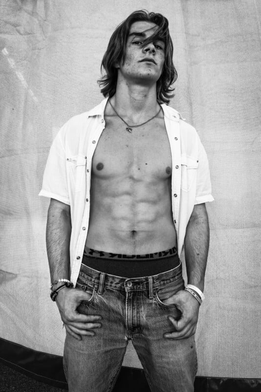 A monochrome photograph featuring a man with long hair standing confidently against a fabric backdrop. He sports an unbuttoned shirt revealing his toned stomach and jeans, with his hands in his pockets and a composed expression.