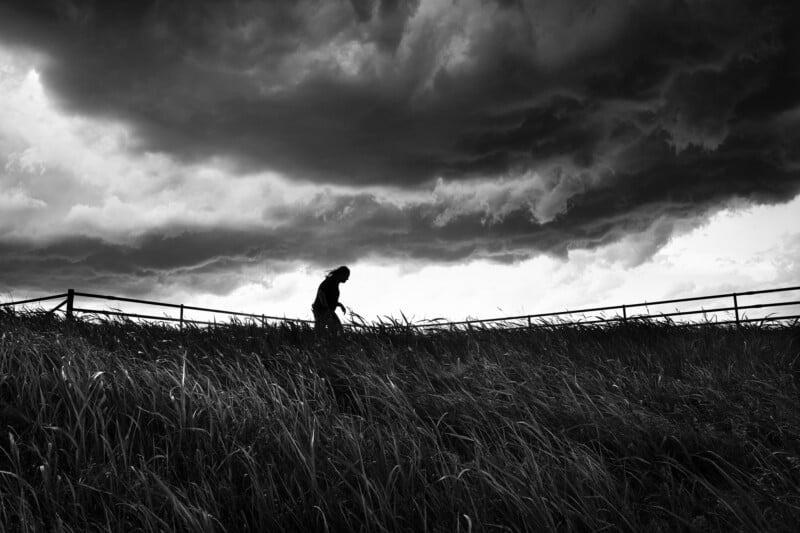 A silhouetted person walks along a grassy hill under a dramatic, cloud-filled sky. The scene is captured in black and white, highlighting the contrast between the dark clouds and the landscape.