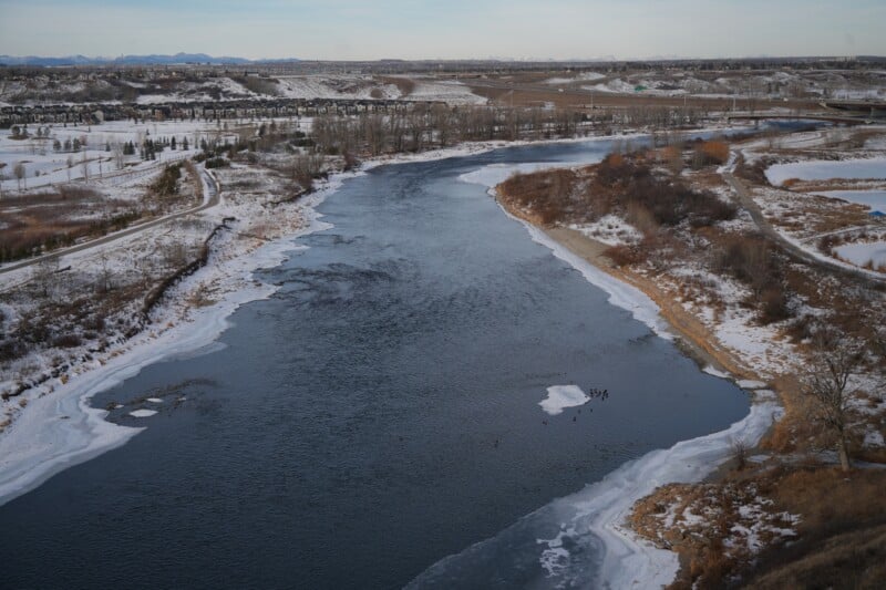 Aerial view of a wide river flowing through a snowy landscape. The river is partially iced over near the banks, surrounded by barren trees and brown, snowy fields stretching into the distance under a cloudy sky.