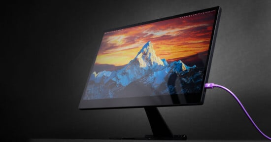 A sleek computer monitor displaying a stunning mountain landscape with a vibrant sunset. The monitor is angled on a minimalist stand against a dark background, with a purple cable connected to it.