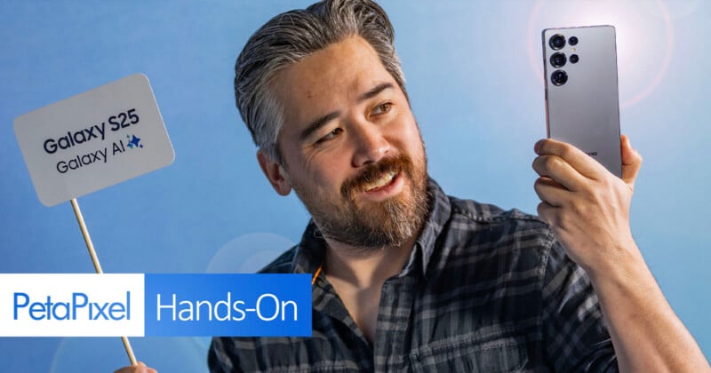 A person with a beard holds up a smartphone, smiling and facing the device. They stand against a blue backdrop. A sign next to them reads "Galaxy S25 Galaxy AI" and "PetaPixel Hands-On.