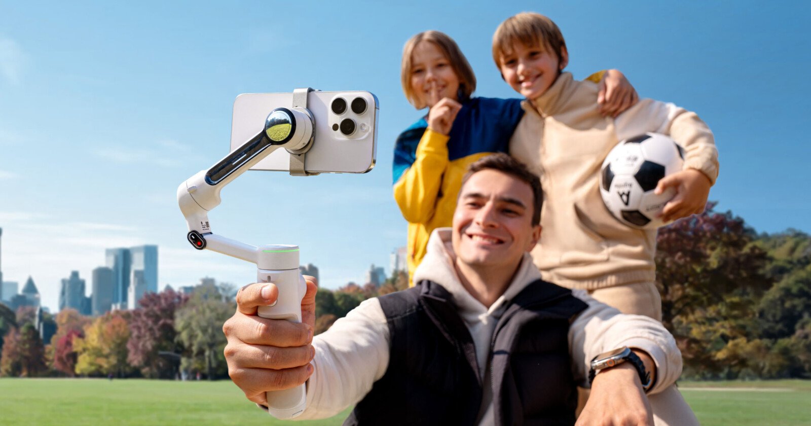 A man uses a smartphone on a stabilizer to take a selfie with two smiling children. The boy holds a soccer ball. They're outdoors in a park with city buildings in the background.