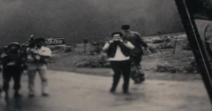 A black and white photograph showing a group of people outdoors. One person is holding a camera, capturing something in the distance. Another is walking away. The background is a grassy landscape under a dark sky.