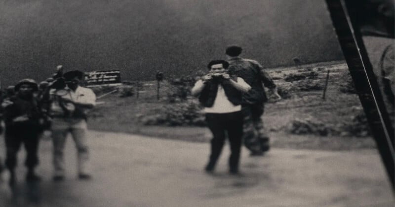 A black and white photo shows a group of people outdoors. Two individuals in focus are looking at cameras, while another person walks away in the background. The scene appears to be candid, with a blurred landscape behind them.