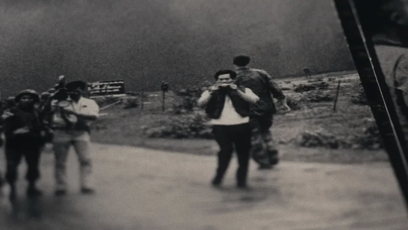 A black and white photograph showing a group of people outdoors. One person is holding a camera, capturing something in the distance. Another is walking away. The background is a grassy landscape under a dark sky.