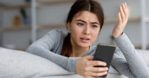 A woman with long brown hair holds a smartphone in one hand while looking confused and frustrated. She is wearing a gray long-sleeve shirt and sitting on a couch with a blurred background of shelves.