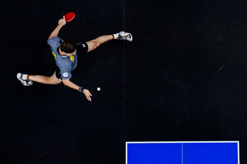 An overhead view of a table tennis player in action, clad in a gray shirt and shorts. The player extends one leg and arm, poised to strike a white ping pong ball with a red paddle. The blue table is partially visible on the right.