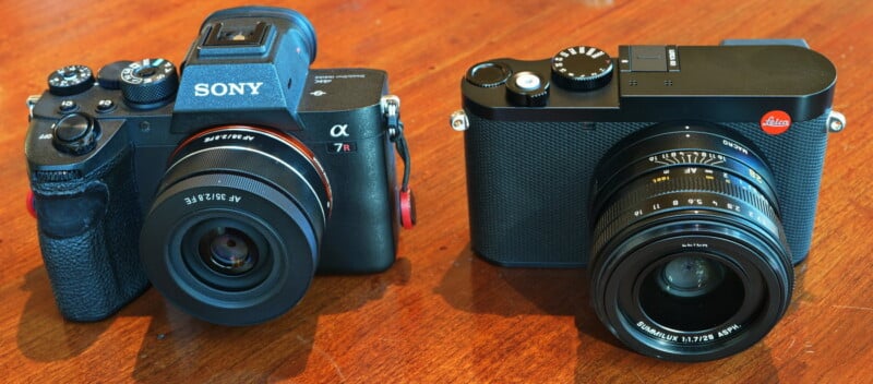 Two cameras on a wooden surface: a Sony Alpha camera on the left with a prominent lens, and a Leica camera on the right, both showcasing their distinct designs and lenses.