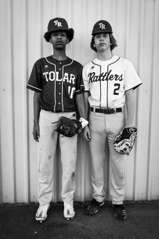 Two youthful baseball athletes stand together in uniform against a metallic wall. One sports a "Tolar" jersey, while the other wears "Rattlers." Both are holding baseball mitts and wearing helmets, gazing at the camera with a grave demeanor. Black and white visual.