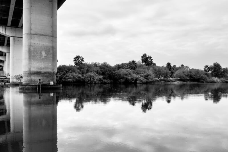 Monochrome photograph of an individual angling beneath a large bridge near a tranquil river. Trees and palm trees line the opposite shore, reflected in the surface of the water. Overcast sky overhead.