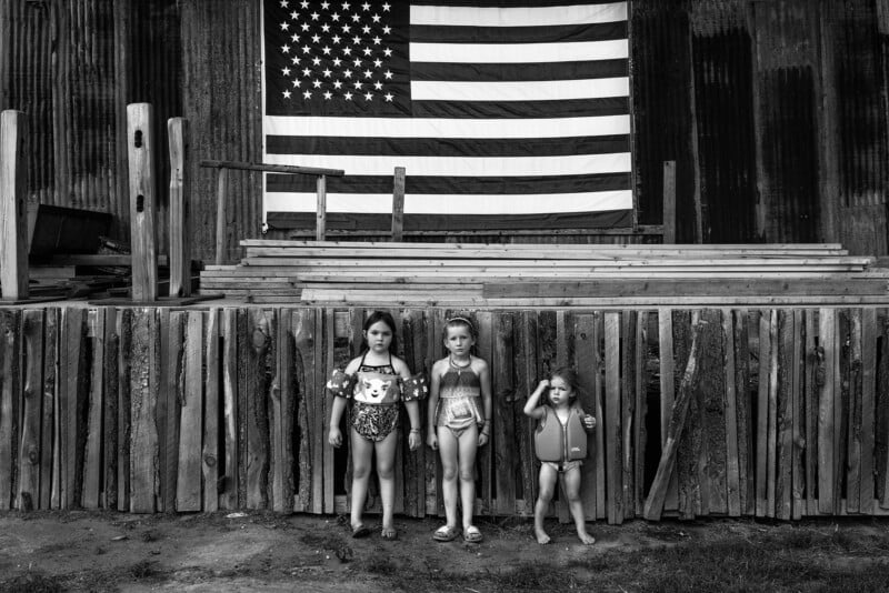 Three children in swimsuits stand in front of a wooden platform with stacks of wood. An American flag is displayed on the structure behind them. The scene is in black and white.