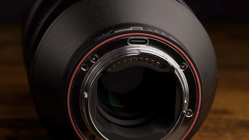 Close-up view of the rear mount of a camera lens, highlighting the metal contacts and circular opening. The outer ring has a red band, and the background is dark, emphasizing the lens details.