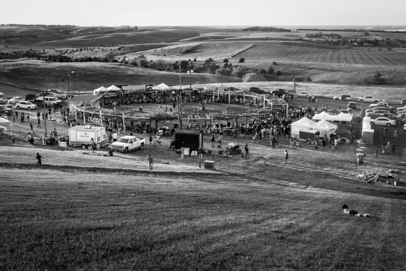 A black and white image of an outdoor gathering on a grassy hill, featuring a large round arena surrounded by people and various tents. Cars are parked nearby, with vast fields and rolling hills in the background.