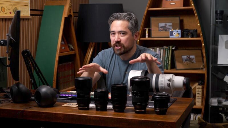 A person with short, gray hair and a beard is sitting at a wooden table indoors. They are gesturing with their hands toward a row of camera lenses in front of them. Shelves with books and photos are visible in the background.