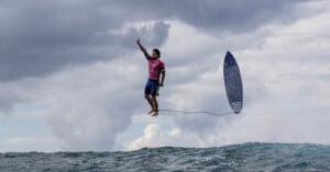 A surfer mid-air above a wave, with a surfboard trailing behind connected by a leash. The person is wearing a pink shirt and blue shorts, pointing upward with one hand. The sky is cloudy, with large waves visible below.