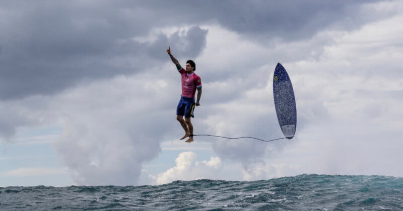 A surfer in mid-air above a wave, with a surfboard trailing behind secured by a leash. The individual is dressed in a pink shirt and blue shorts, pointing upwards with one hand. The sky appears cloudy, showcasing large waves below.