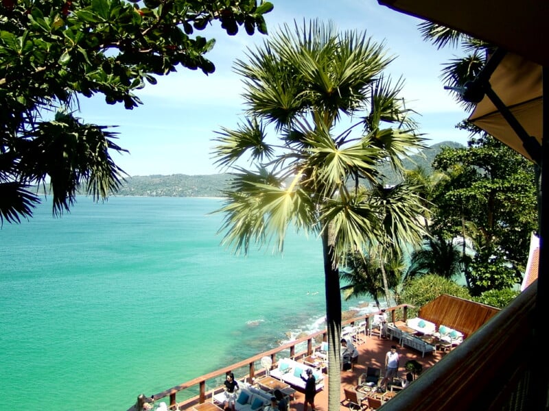 Scenic view of a coastal area with turquoise water and palm trees. A terrace with tables is set up overlooking the ocean. People are gathered around the tables enjoying the view. The backdrop includes lush greenery and hills.