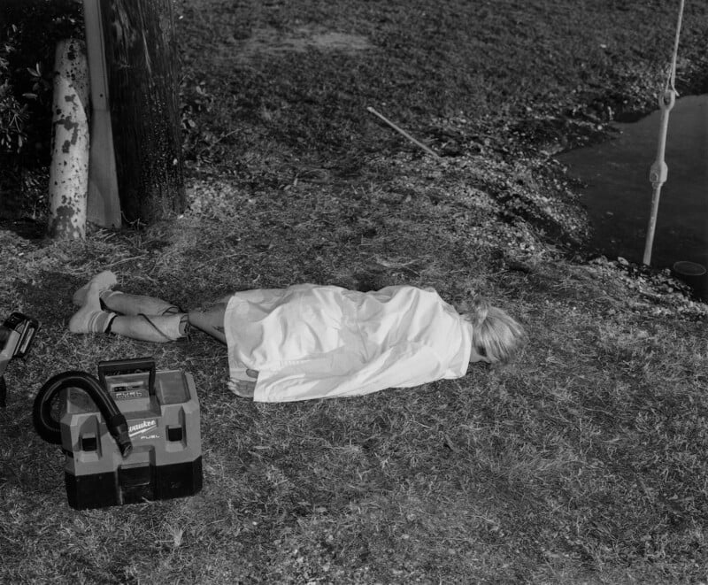 A black and white image of a person lying face down outdoors on grass, covered in a sheet. Nearby is a vacuum cleaner and a tree trunk. A water body or pond is visible to the right. Scene appears serene yet enigmatic.