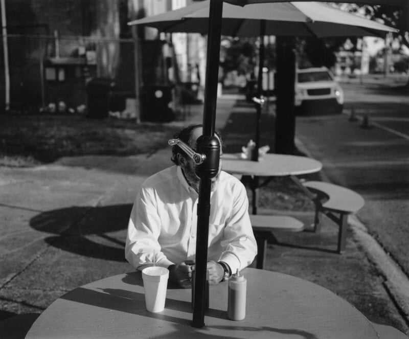 A person in a white shirt sits at an outdoor table with an umbrella post obscuring their face. On the table are a cup and a condiment bottle. The scene is in black and white, capturing a sunny street setting with benches and a car in the background.